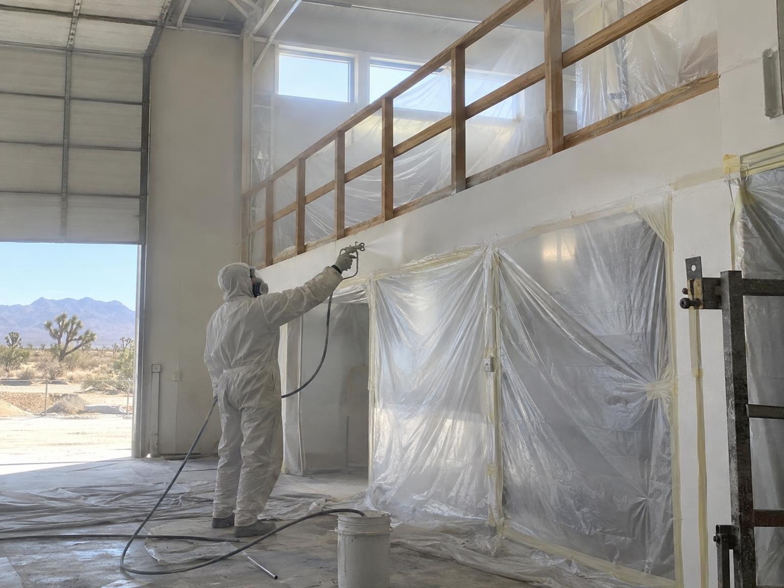 Painter in coveralls and a respirator spraying a commercial mezzanine behind plastic masking